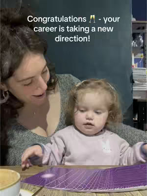 A woman with dark curly hair is seen from the chest up, holding a young child. The child, with light-colored curly hair, is looking down at a deck of tarot cards on a table. The woman is looking at the child. The background is a dark wall with some items on a shelf.