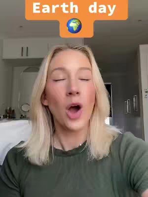 A selfie-style video cover featuring a young woman with blonde hair and makeup. Above her head floats an orange banner with white text that reads 'Earth day', and a globe graphic. Her mouth is open, and her eyes are closed, suggesting she's singing or speaking with emphasis. The background is soft-focus, featuring a room with closed cabinets and basic decor. The color palette is natural, with the woman's green top contrasting the neutral tones of the room and banner.