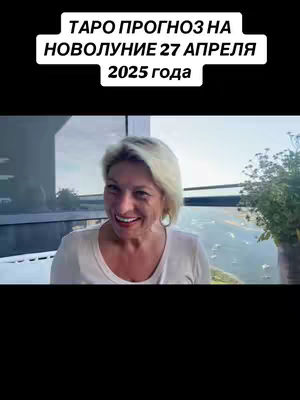 A woman with short blonde hair smiles widely, with a slight tilt of her head. She is wearing a white or light-colored top. Behind her is a view of a cityscape.