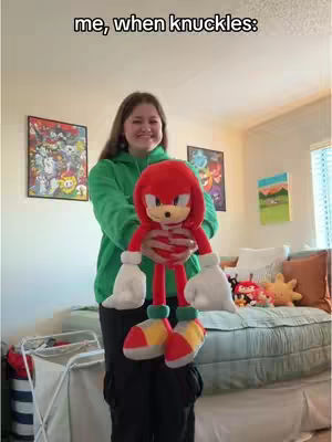 A young woman stands in a room, holding a large Knuckles the Echidna plush toy. She's grinning, and the plush is in front of her. The background suggests a bedroom with a bed and some wall decorations.