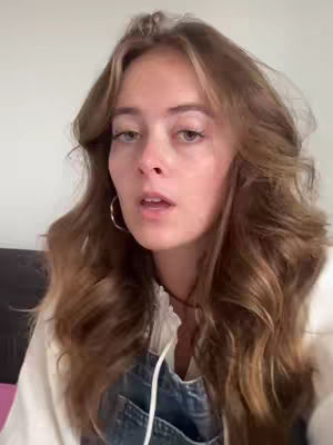 A young woman with long, wavy brown hair and a white top is looking towards the camera. She is wearing a pair of hoop earrings. The background is a plain, light-colored wall. The composition is a medium close-up, focusing on her face and upper body.