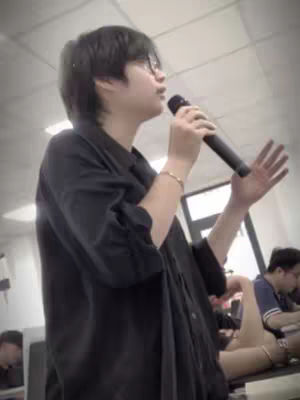 A person with short black hair, wearing glasses and a black shirt, is holding a microphone with one hand and gesturing with the other. The background is a blurred interior space, likely an office or meeting room. Other people are also present in the background.
