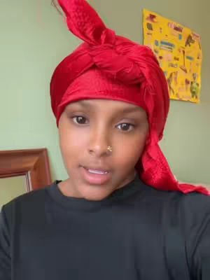A young woman with a soft complexion is the focus of the image, framed from the chest up. She is looking directly at the camera, positioned slightly off-center. Her dark hair is completely covered by a vibrant red fabric head wrap, knotted at the top. She has a small nose ring. She is wearing a plain black top. The background is simple, featuring a green-painted wall along with a glimpse of a picture hanging. The lighting appears natural and soft, highlighting her face and the color contrast of her attire. The overall composition is straightforward, drawing attention to the subject.