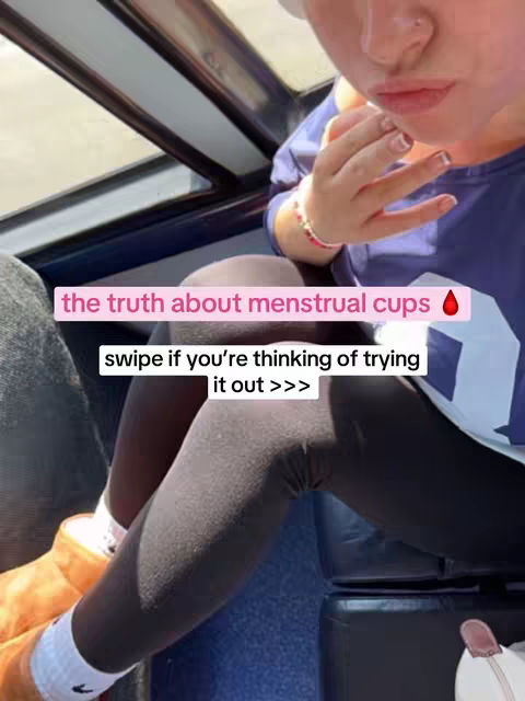 A selfie-style shot featuring a person's face and upper body, with the focus on their mouth and hand gesture. They touch their lips with their fingers, and a pink text box overlays the image, with the words 'the truth about menstrual cups' and a drop of blood-shaped emoji. Further text below reads 'swipe if you're thinking of trying it out >>>'. The person wears a blue top and black leggings, suggesting a casual setting, possibly indoors, with a glimpse of a seat and window in the background.