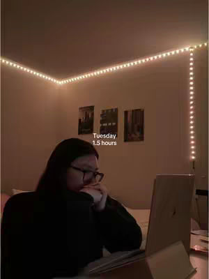 A person sits at a desk in a dimly lit room, illuminated primarily by string lights. The person has dark hair, wears glasses, and is facing a laptop. On the wall behind them are framed pictures. The composition is close-up, focusing on the person and the workspace.