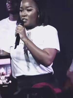 A person, presumably a vocalist, is facing the camera and holding a dark microphone. They wear a white t-shirt and black bottoms. Partially visible behind them, a person in a white shirt is out of focus, and a dark background gives the photo a concert hall vibe.