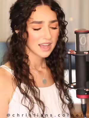 A woman with long, wavy dark hair is singing into a microphone. She is wearing a white top and a necklace. The background is blurred, but appears to be a room with a blue wall.