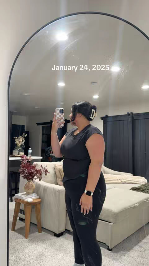 A woman stands in front of a large arched mirror, taking a photo of herself. She wears a black top and leggings, and holds a phone, with the date in white text on the mirror. The background reveals a modern living space with a neutral color palette.
