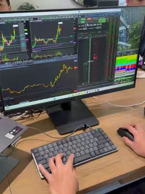 The image shows a person's hands using a keyboard and mouse in front of a large computer monitor displaying stock market graphs and data. A second monitor is to the right, and the background setting is blurred. The scene suggests a focus on day trading or financial analysis.
