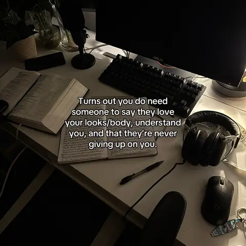 A top-down view of a desk setup. A desktop computer with a matte black monitor. A black keyboard and mouse are on the desk. A journal is open to a handwritten page with dark pen and various supplies around it, like headphones and a microphone.