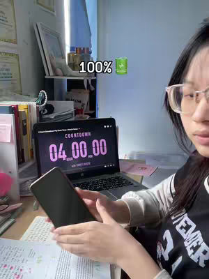 A young woman with black hair and glasses is seated at a desk. She is holding a smartphone in her hands, while looking towards the right corner, with a laptop open in front of her displaying a countdown timer. Various items such as books, papers, and small decorative objects are visible.