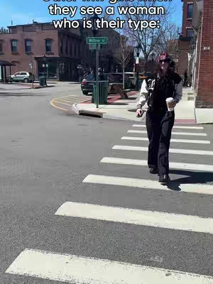 The image shows a woman walking across a crosswalk on a sunny day. She is holding a coffee cup and is wearing sunglasses, a jacket with a letter "T" on it and dark pants. The background includes buildings, street signs, and a crosswalk. The overall composition is a candid shot capturing a moment in an urban setting.