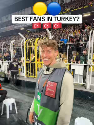 A man wearing a vest stands in front of a crowd at a sports stadium. The man is smiling and looking at the camera. Above him, there is a white text box with the words "BEST FANS IN TURKEY?" and two circle emojis and three Turkish flags emojis.