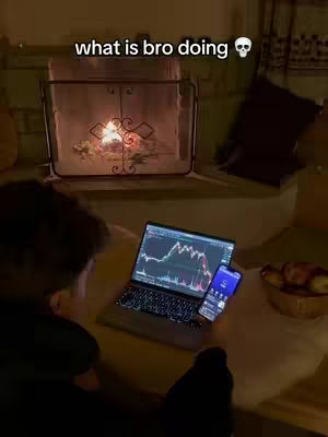A person is sitting at a table with a laptop and smartphone. The laptop is displaying a stock chart, and the smartphone is also on the table. Behind them is a fireplace with a lit fire. The overall setting is indoors and dimly lit.