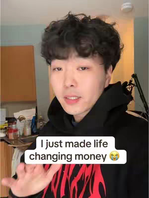 A young Asian man with curly black hair is in a selfie video. He wears a black hoodie. A white rectangular text box overlays the bottom of the frame, with text in white lettering and an emoji. The background is a blurred interior setting.