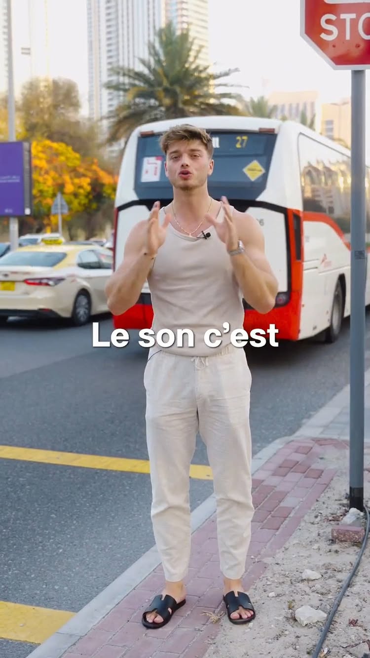 A man in a tank top and slacks stands on a city street, gesturing with both hands, with a bus and buildings in the background. The text overlay, "Le son c'est", is visible.