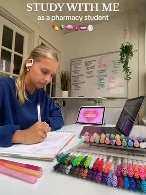 A young woman is captured in a well-lit room, likely her apartment. She is seated at a desk, intently focused on writing in a notebook. Various study materials and technological devices surround her, including a laptop displaying the time, colored markers, and a tablet showing a clock, all of which suggests a study or productivity setting. The background features a clean white wall, a window, and minimal decorative elements.