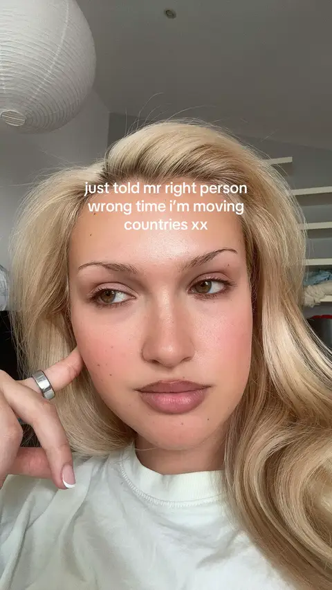 A selfie of a young woman with long blonde hair, fair skin, and makeup looking at the camera in close-up. She is wearing a white t-shirt and a silver ring. Text overlayed on the image reads, "just told mr right person wrong time i'm moving countries xx". The background is out of focus and appears to be an interior setting.
