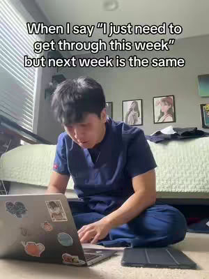 A young man wearing scrubs sits on the floor with a laptop open in front of him, appearing to be studying or working. The setting is a bedroom with a bed visible in the background, a window with blinds, and artwork on the wall. Additional items include a tablet and stickers on the laptop. The overall composition gives a sense of a casual, study-related environment.