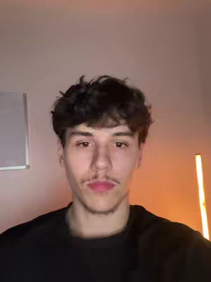 A selfie-style video featuring a young man with dark curly hair and a slight mustache, seen from the chest up. He's centered in the frame, looking directly at the viewer with a neutral expression. The background is softly lit with warm tones on the right side, which contrasts with the cooler, muted tones on the left. His attire is a simple black shirt that focuses attention on his face.