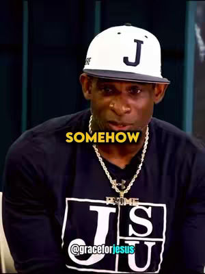 A man wearing a white cap with a black 'J' on it with a black shirt and necklace. The text overlay says 'SOMEHOW' in yellow letters.