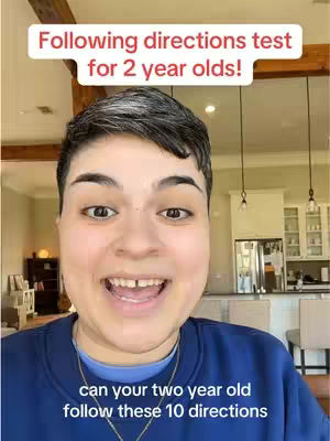 A selfie-style video cover image features a woman with short, dark hair smiling directly at the camera. The woman is wearing a blue sweatshirt and a gold necklace. Two text overlays are present: a white rectangular box near the top with the text 'Following directions test for 2 year olds!' and a smaller white box towards the bottom with the text 'can your two year old follow these 10 directions.' The background includes a living room area with a kitchen visible, suggesting an indoor setting.