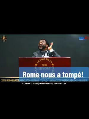A man speaks from behind a podium with a banner that reads "Rome nous a trompé!" (Rome deceived us!) in front of a dark background, The man is wearing a black suit with a bow tie. The foreground features the speaker, a podium displaying text and logos, and a sign with text, and background is a dark setting.