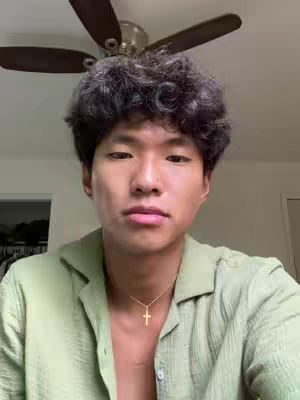 A face-close up of a young Asian man is framed from the chest up. He has short, dark curly hair, and wears a light green, button-down shirt, slightly unbuttoned at the top, revealing a gold cross necklace. His facial expression is neutral. Above him, a ceiling fan is partially visible. The background is softly blurred with neutral tones, and the lighting appears natural.