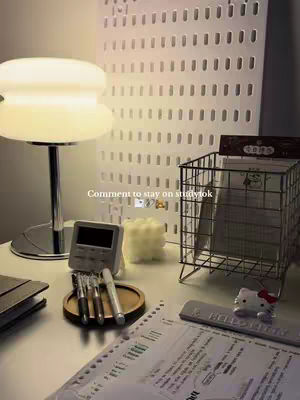 A well-lit desk setup featuring a white lamp, a digital clock, stationary, and papers. A metal wire basket, a Hello Kitty figurine, and a white, perforated wall panel are also included.