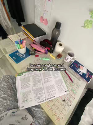 The image shows a study desk with open books, stationery, a mug, and other study-related items. The text overlay reads 'Become a ghost and improve in the silence'.