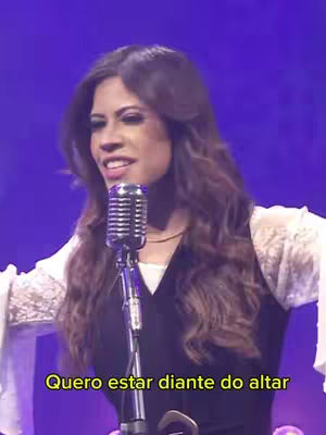 A medium shot features a woman with long brown hair, holding a vintage microphone in front of her. She is wearing a black vest over a white, lacy top. The background is a blurred purple hue. Text overlayed at the bottom reads 'Quero estar diante do altar' in a yellow font.