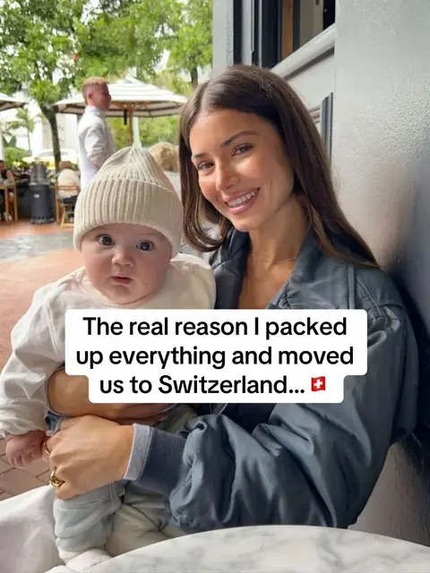 A woman holds a baby, both smiling at the camera. The woman has long brown hair, and wears a casual outfit and a light jacket. Behind her, there is a blurred outdoor or restaurant setting with trees.