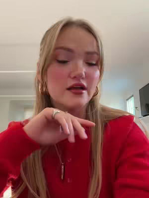 A young woman with long blonde hair and makeup looks slightly off-camera, wearing a red sweater. Her hand is up near her face as she speaks, and the background suggests a well-lit, neutral-toned interior.