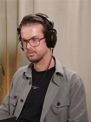 A man wearing glasses and headphones in an indoor studio shot. The man seems to be talking as shown by the close up shot. The background seems to be a beige wall.