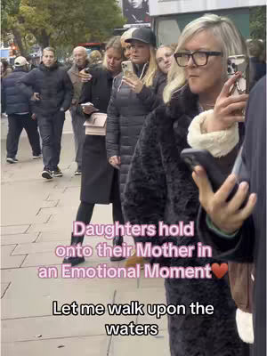 The image shows a group of people on a city sidewalk. Several people are holding smartphones, with one woman in the foreground holding her phone up to film. Text overlays read 'Daughters hold onto their Mother in an Emotional Moment' and 'Let me walk upon the waters'. The overall scene is shot during the day, with a focus on the people and their expressions.