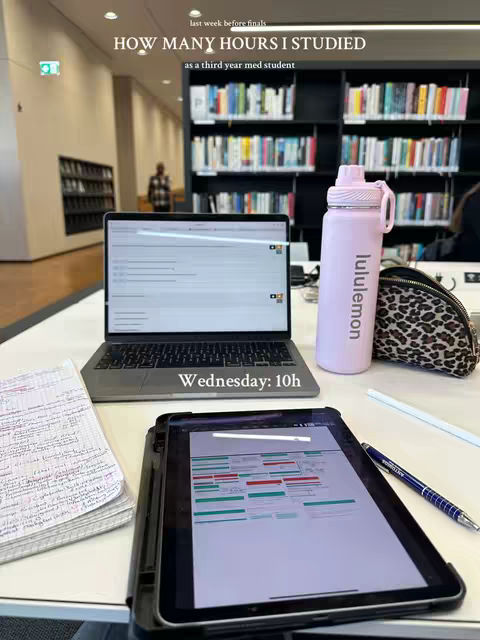 A desk in a library is shown, on which a laptop, a tablet, a pink water bottle, and a notepad are placed. The laptop displays a screen with a study schedule. The tablet shows a similar schedule, and the water bottle has the brand 'lululemon'. Handwritten notes are sitting next to the notepad. In the background, there are rows of bookshelves and a person walking in the distance.
