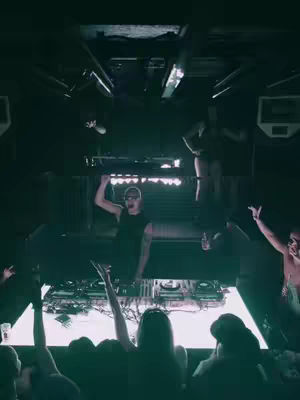 The image captures an energetic concert atmosphere. The perspective is from below, looking up at a stage where a DJ is performing, and a crowd is visible at the bottom. The lighting is dim with strong spotlights creating contrast. The DJ, a man wearing glasses, is gesturing with one hand raised. In the background, other individuals are captured in motion, dancing or interacting with the music. 