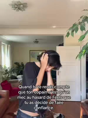 The image shows a person indoors in a home setting, possibly a living room. The person, who seems to be a woman, is the central focus, with their face hidden behind their hands. She has short dark hair. They are wearing a black short sleeve shirt and blue jeans. Text overlayed on the image provides context to the scene's emotion, which seems to revolve about feelings of dismay.