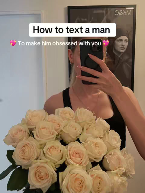 A woman is holding a bouquet of cream-colored roses in front of a mirror. She is taking the photo with her phone, which obscures her face. The background has a black framed art piece. The photo has a soft, natural light, and the overall composition is balanced.