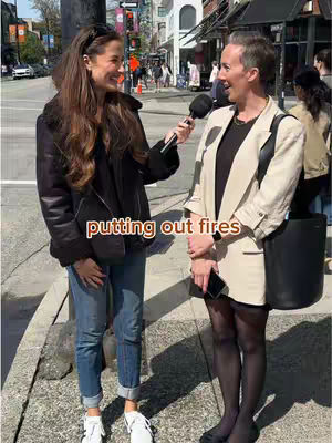 Two women are standing on a city sidewalk, near a street intersection. One holds a microphone and appears to be interviewing the other. The woman with the microphone is wearing a black leather jacket and jeans. The other woman is wearing a white blazer and black pantyhose, holding a phone. The text overlay says: putting out fires.