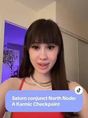 A woman with dark hair and bangs is the focal point of the image. She has fair skin, is wearing a red top and a pearl necklace, and is looking directly at the camera with a neutral expression. A text overlay in a light blue rounded rectangle appears at the bottom, it reads "Saturn conjunct North Node: A Karmic Checkpoint". A hint of a door frame with a soft, lavender hue and a small tree with lights provides the interior setting. A subtle TikTok logo is noticeable.