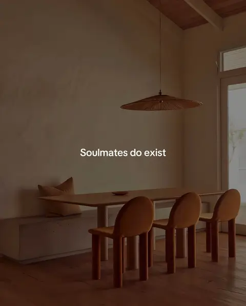 A well-lit interior dining scene featuring a wooden table with three rounded chairs, a cushioned bench, and a hanging wicker lamp. The walls are a neutral color, setting a calm and comforting mood. The text overlay states 'Soulmates do exist'.