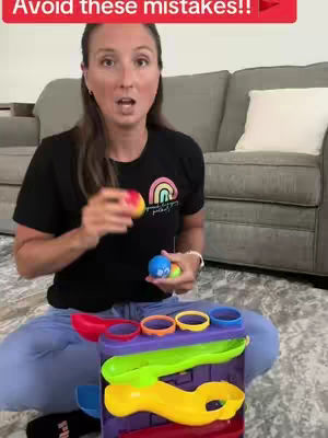 A woman in a black t-shirt is holding colorful toy balls over a multi-colored plastic toy. She appears to be talking to the camera, with a gray couch and white pillow in the background.