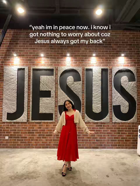 A woman in a red dress stands in front of a brick wall with the word 'JESUS' displayed in large letters. She has her arms outstretched, and the image conveys a sense of peace and faith.