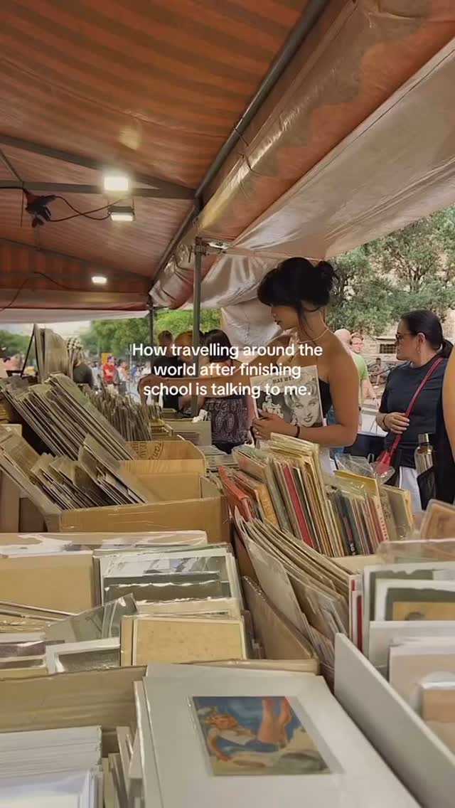 A woman browsing items at an outdoor market, with a blurred background of other people shopping. The image is warm-toned and the composition is focused on the items for sale and her interaction with them.