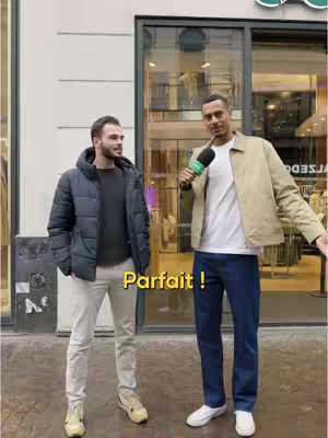 Two men stand in front of a shop entrance, one holding a microphone. A yellow text overlay reads 'Parfait!'