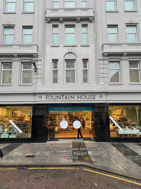 The image shows the front of a Primark Home store in a multi-story building. The store has large glass windows displaying merchandise. The text overlay says 'Primark home is open tomorrow. Let's see what they got Belfast'. The storefront is below a building facade of repeating windows.