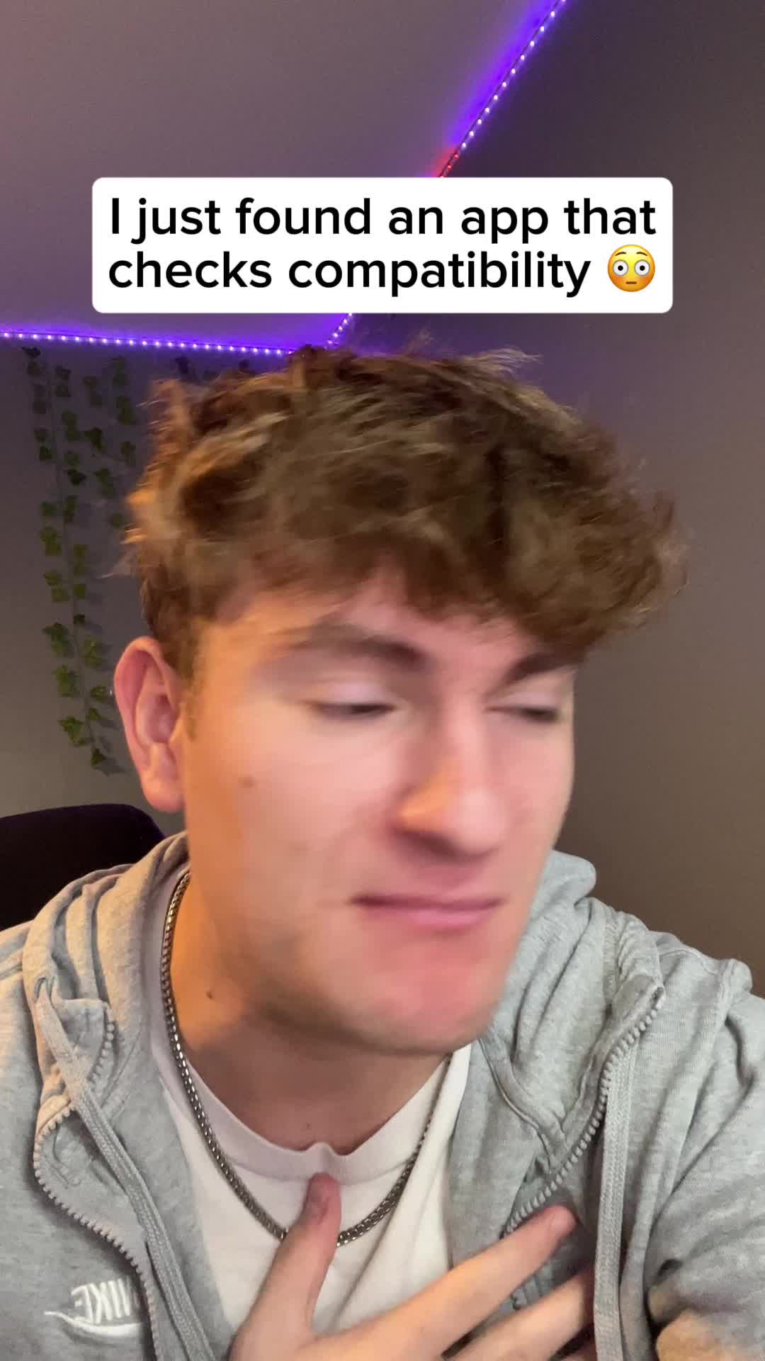 A young man with light brown hair and a gray hoodie is centered in the frame, looking slightly to the side with a slightly surprised expression. A text overlay in a white box reads, 'I just found an app that checks compatibility' with an emoji. The background is a blurred room lit with soft lighting.