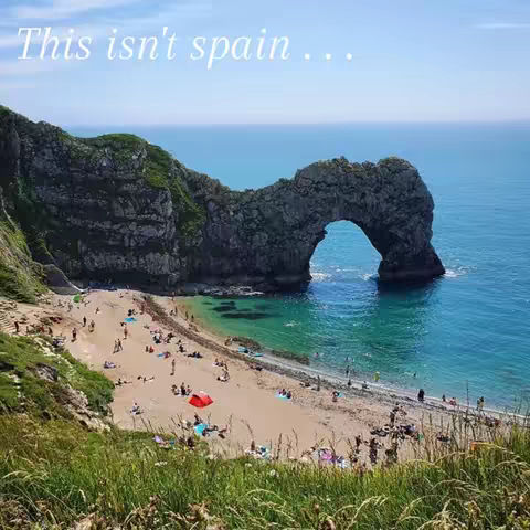 An aerial view shows a picturesque coastal scene. A large, natural stone arch spans the turquoise water. A sandy beach is dotted with sunbathers and colorful umbrellas, backed by green cliffs on the left. The sky is clear and light blue.