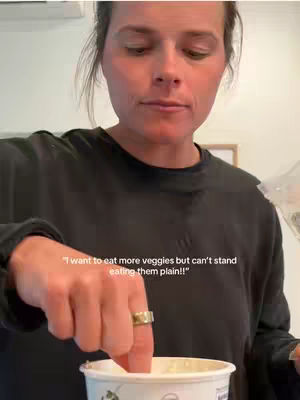A woman is shown in a selfie, appearing from the chest up, dipping a finger into a container of white dip. Overlaid text mentions eating veggies, and the overall look is casual domestic.